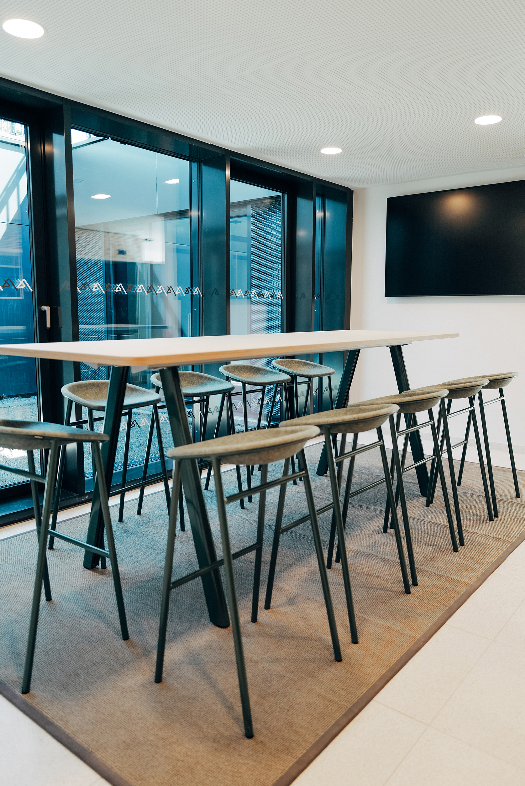 Flexibler Besprechungsbereich mit langem Tisch und Sitzhockern vor einer verglasten Raumfassade